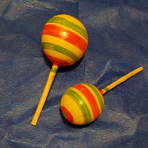 Könnte beinhalten: Zwei Holzmaracas mit gelben, roten und grünen Streifenmustern. Die Maracas haben lange, dünne Holzgriffe. Der Hintergrund ist eine strukturierte blaue Oberfläche.