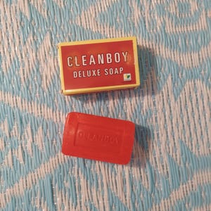 May include: A red bar of soap with the words "Cleanboy Deluxe Soap" printed on it. The soap is sitting on a blue and white patterned surface.