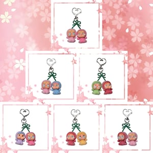 May include: Keychains featuring pairs of small, stylized figures with heart-shaped eyes and hooded outfits. Each keychain has a silver-colored ring and a green decorative element. The figures come in various color combinations, including pink, blue, green, and red.