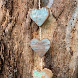 May include: Three wooden hearts hanging from a rope, painted in shades of blue and green with a distressed finish. The hearts are hanging against a tree trunk.