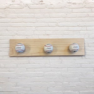 May include: A wooden coat rack featuring three baseballs. Each baseball is white with red stitching and the words "OFFICIAL LEAGUE" are printed on them. The rack is mounted on a white brick wall.