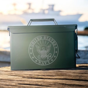 May include: An olive green metal box with a handle and a hinged lid. The front of the box features the words "UNITED STATES NAVY" surrounding an eagle emblem. The box is sitting on a wooden surface.