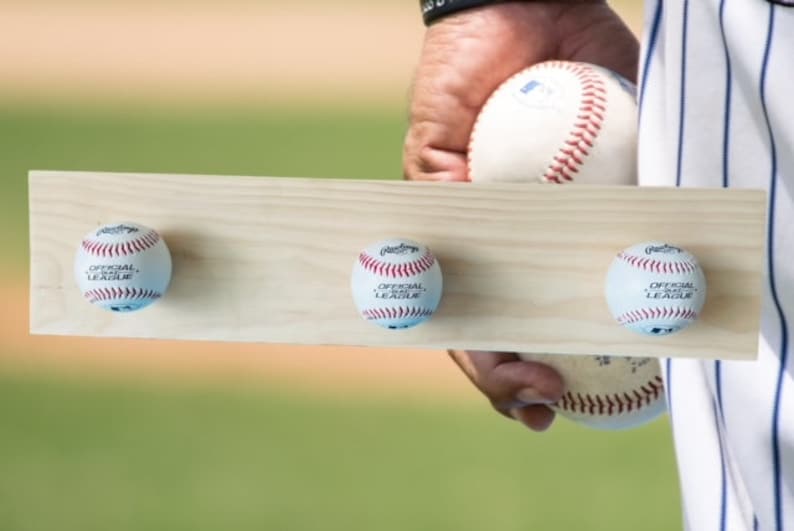 May include: A wooden baseball display board with three baseballs attached. The baseballs are white with red stitching and the words "OFFICIAL LEAGUE" printed on them. The board is held by a person.