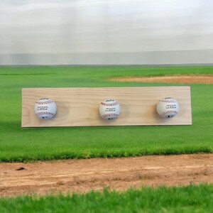 May include: A wooden baseball display featuring three baseballs. Each baseball is white with red stitching and the text "OFFICIAL LEAGUE". The wooden board is light brown and has three circular cutouts to hold the baseballs.