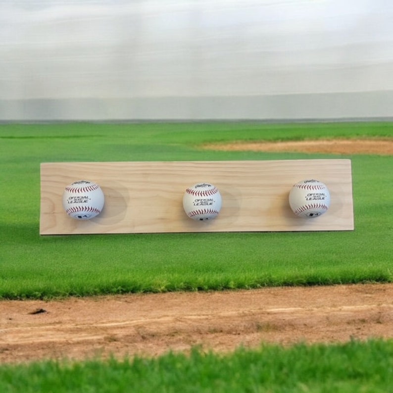 May include: A wooden baseball display featuring three baseballs. Each baseball is white with red stitching and the text "OFFICIAL LEAGUE". The wooden board is light brown and has three circular cutouts to hold the baseballs.