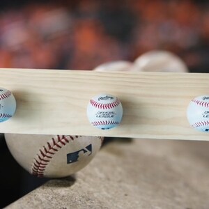 May include: A wooden baseball-themed coat rack featuring three baseballs attached to a light-colored wooden plank. Each baseball is white with red stitching and the words "OFFICIAL LEAGUE" printed on it. A baseball rests below the rack.