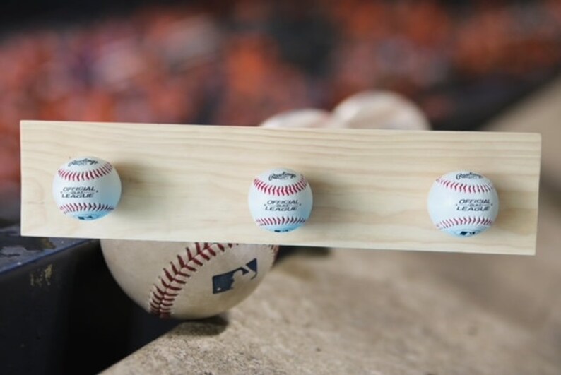 May include: A wooden baseball-themed coat rack featuring three baseballs attached to a light-colored wooden plank. Each baseball is white with red stitching and the words "OFFICIAL LEAGUE" printed on it. A baseball rests below the rack.