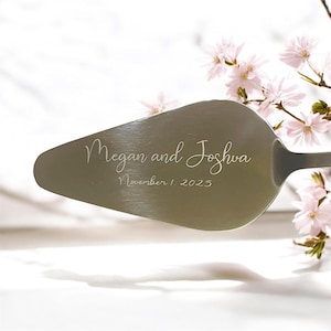 Puede incluir: Una espátula plateada para pastel con los nombres grabados "Megan and Joshua" y la fecha "November 1, 2023". La espátula se muestra con delicadas flores rosas y un fondo blanco.