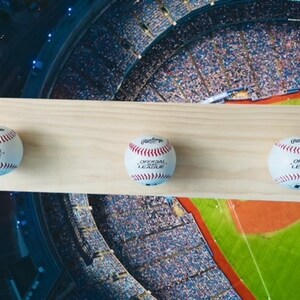 May include: A wooden baseball display shelf with three baseballs. Each baseball is white with red stitching and the words "OFFICIAL LEAGUE" printed on it. The shelf is made of light-colored wood.