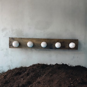 May include: A wooden baseball-themed coat rack with five baseballs attached. The baseballs are white with red stitching. The wood is dark brown and mounted on a gray wall. The rack is a decorative item.