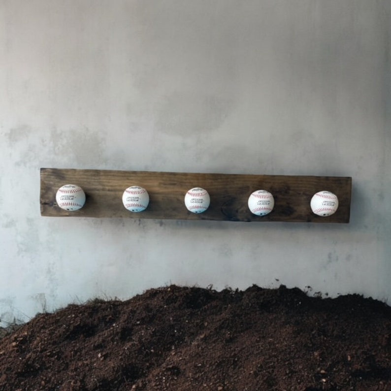 May include: A wooden baseball-themed coat rack with five baseballs attached. The baseballs are white with red stitching. The wood is dark brown and mounted on a gray wall. The rack is a decorative item.