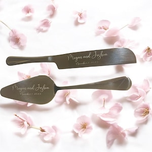 Puede incluir: Un juego de cuchillo y paleta para pastel plateados con los nombres "Megan and Joshua" y la fecha "November 1, 2025" grabados. El juego se exhibe sobre una superficie blanca con pétalos de flores rosas esparcidos.