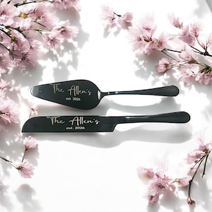 Puede incluir: Juego de servidor y cuchillo para pastel negros con la inscripción "The Allen's est. 2026". El juego se muestra sobre una superficie blanca, rodeado de ramas de flores de cerezo rosas. El servidor tiene una hoja ancha y plana, y el cuchillo tiene un borde dentado.