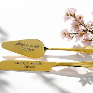 Puede incluir: Un juego dorado de cuchillo y paleta para pastel, personalizado con los nombres "Patrick & Michelle Johnson" y la fecha "04.27.2024". El juego está sobre una superficie blanca, con flores rosas en el fondo.