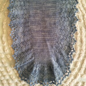 May include: A gray knitted shawl with a crocheted lace border. The shawl is made with a gradient yarn that transitions from a light gray to a darker gray. The shawl is shown on a white background.