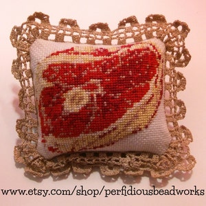 May include: A small, white pillow with a crocheted lace trim. The pillow features a red and white embroidered image of a steak.