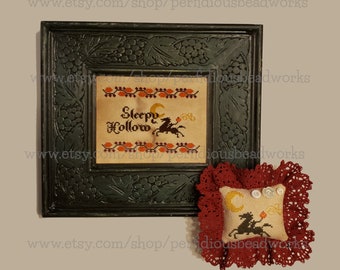 May include: A framed cross-stitch featuring the words "Sleepy Hollow" with a silhouette of a rider and horse, a crescent moon, and pumpkins. A small, matching pin cushion with a red lace border is also present.