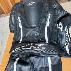 Op de afbeelding: Zwart leren motorracepak met witte accenten en het Alpinestars-logo. Het pak heeft een rugprotector en kniebeschermers.