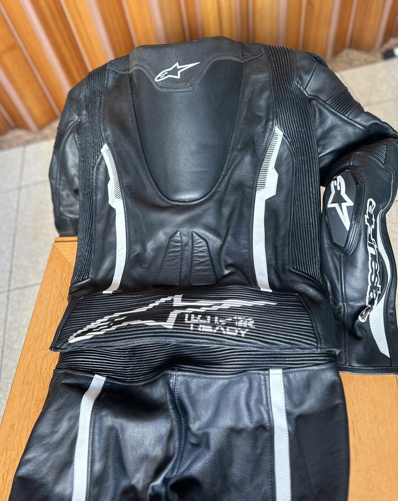 Op de afbeelding: Zwart leren motorracepak met witte accenten en het Alpinestars-logo. Het pak heeft een rugprotector en kniebeschermers.