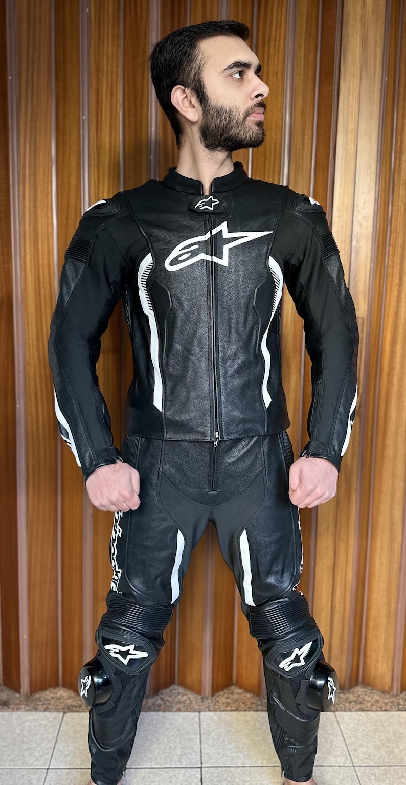 Op de afbeelding: Zwart leren motorpak met witte accenten en een sterlogo. Het pak heeft kniebeschermers en een ritssluiting.