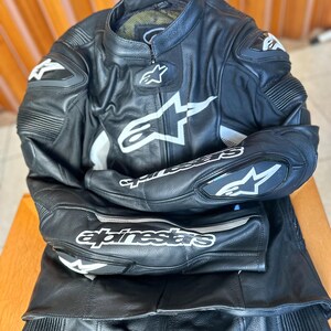 Op de afbeelding: Zwart leren motorpak met witte en zilveren accenten. Het pak heeft het Alpinestars logo en branding.
