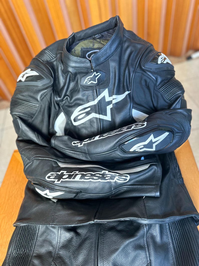 Op de afbeelding: Zwart leren motorpak met witte en zilveren accenten. Het pak heeft het Alpinestars logo en branding.