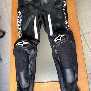 Op de afbeelding: Zwarte leren motorbroek met witte accenten en kniebeschermers. De broek heeft de tekst "Alpinestars" en "S-Sport" op de benen.