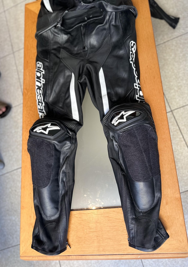 Op de afbeelding: Zwarte leren motorbroek met witte accenten en kniebeschermers. De broek heeft de tekst "Alpinestars" en "S-Sport" op de benen.