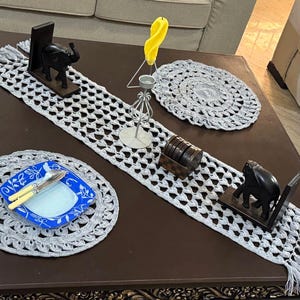 Könnte beinhalten: Ein grauer gehäkelter Tischläufer mit Fransen an den Enden. Der Läufer liegt auf einem dunkelbraunen Tisch mit zwei hölzernen Elefanten-Buchstützen, einem kleinen grauen gehäkelten Untersetzer und einem blauen und weißen Keramikteller mit zwei silbernen Messern.