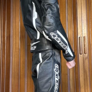 Op de afbeelding: Een zwart leren motorpak met witte accenten en het woord "alpinestars" gedrukt op het been. Het pak heeft een rits aan de voorkant en een riem om de taille.