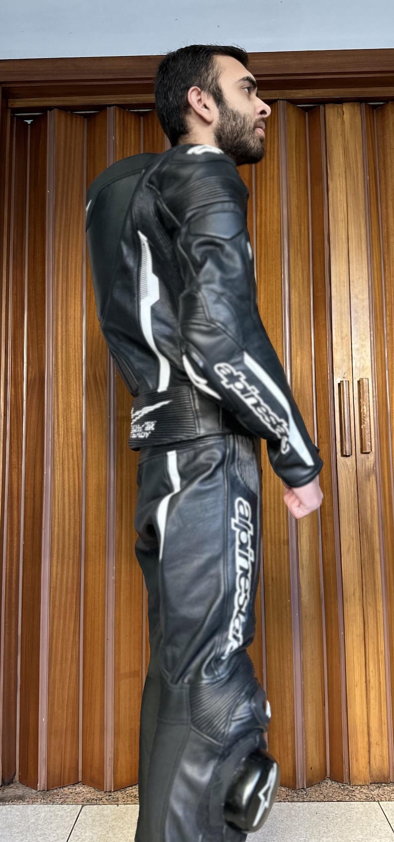 Op de afbeelding: Een zwart leren motorpak met witte accenten en het woord "alpinestars" gedrukt op het been. Het pak heeft een rits aan de voorkant en een riem om de taille.