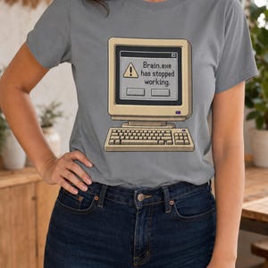 May include: A grey short-sleeved t-shirt featuring a graphic of a vintage beige computer monitor and keyboard. The screen displays a classic error window with a yellow warning icon and the text, Brain.exe has stopped working.