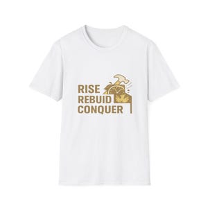 Könnte beinhalten: Ein weißes T-Shirt mit kurzen Ärmeln und einem beigefarbenen und braunen Aufdruck. Das Motiv zeigt den Text "RISE REBUILD CONQUER" sowie eine Illustration eines Hammers, eines Zifferblatts und eines Phönix, der aus Flammen aufsteigt.