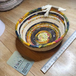 May include: A handcrafted, circular basket with a spiral design. The basket has a beige woven base with colourful fabric strips in yellow, green, and blue. A white rope handle is attached to the rim. A ruler is next to the basket.