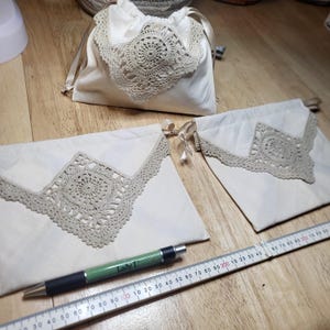 May include: Off-white drawstring bags with beige crochet lace trim. One bag is closed, and two are open. A green pen and a ruler are also in the image. The bags are on a wooden surface.