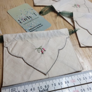 May include: Cream fabric pouch with a scalloped edge, embroidered with pink berries and green leaves. It has a green ribbon drawstring closure. A ruler shows measurements in centimetres. A business card with the text "Lady T" is visible.