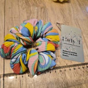 May include: A vibrant scrunchie in shades of pink, yellow, blue, and orange. The scrunchie is placed near a tag with the text "Lady T" and "Making & Creating Whatever My Heart Desires."