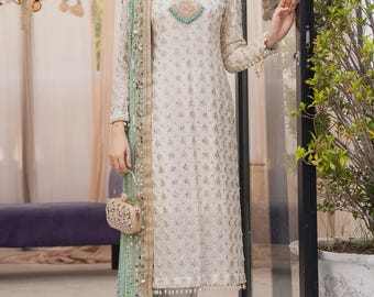 Puede incluir: Un vestido marfil de manga larga con intrincados bordados dorados y una bufanda a juego. El vestido presenta un escote de ojo de cerradura con detalles turquesa. El conjunto incluye un pequeño clutch adornado y sandalias doradas.