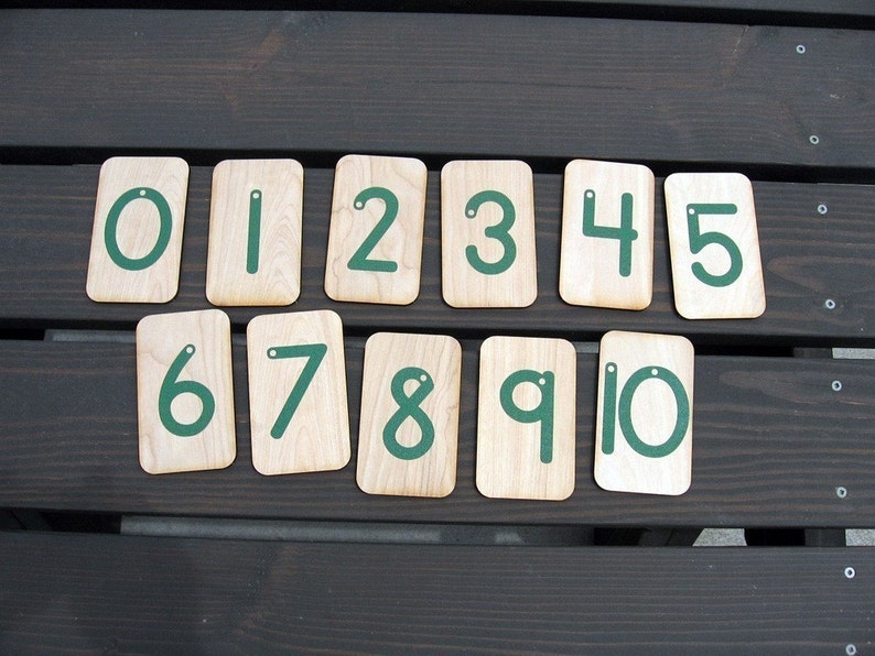 May include: Ten wooden cards with numbers 0 through 10 written in green on them. The cards are arranged in a row on a dark brown wooden surface.