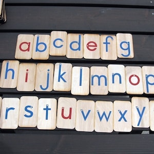 May include: Wooden alphabet letter tiles with blue and red painted letters. The letters are arranged in alphabetical order on a wooden surface.