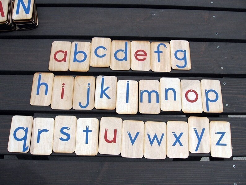May include: Wooden alphabet letter tiles with blue and red painted letters. The letters are arranged in alphabetical order on a wooden surface.