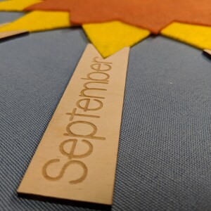 May include: A wooden sign with the word "September" written on it. The sign is a light brown color and has a slightly rounded shape. The sign is sitting on a blue fabric surface.