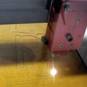 May include: A close-up of a laser cutter cutting a design into a piece of yellow wood. The laser cutter is red and has a small black button on the side. The design being cut is a letter "R".
