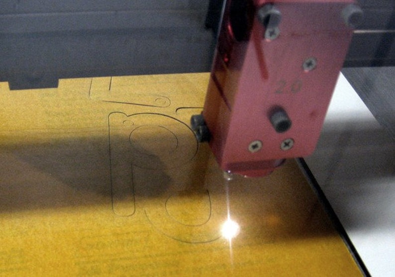 May include: A close-up of a laser cutter cutting a design into a piece of yellow wood. The laser cutter is red and has a small black button on the side. The design being cut is a letter "R".