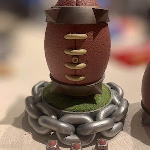 May include: A brown football with a gold chain base and three smaller footballs with gold chains. The footballs are on a light brown surface.