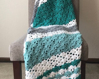 May include: A handmade crochet blanket draped over a gray chair. The blanket features a diagonal pattern of teal, white, and gray stripes. The texture of the crochet stitches is visible, adding to the cozy aesthetic. The blanket is a soft home decor item.