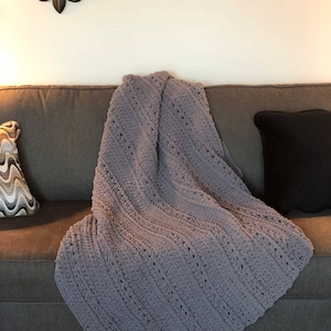 May include: A soft, gray crocheted blanket draped over a gray sofa. The blanket has a textured pattern and is folded diagonally. A decorative pillow with a black pillowcase and a patterned pillow are also on the sofa.