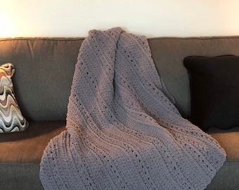 May include: A soft, gray crocheted blanket draped over a gray sofa. The blanket has a textured pattern and is folded diagonally. A decorative pillow with a black pillowcase and a patterned pillow are also on the sofa.