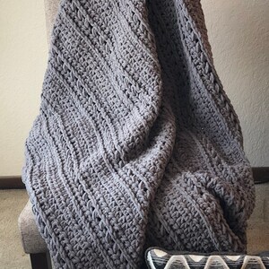 May include: A large, gray, hand-crocheted blanket draped over a gray upholstered chair. The blanket has a thick, textured weave. A decorative pillow with a wavy pattern in shades of brown, gray, and white is visible.