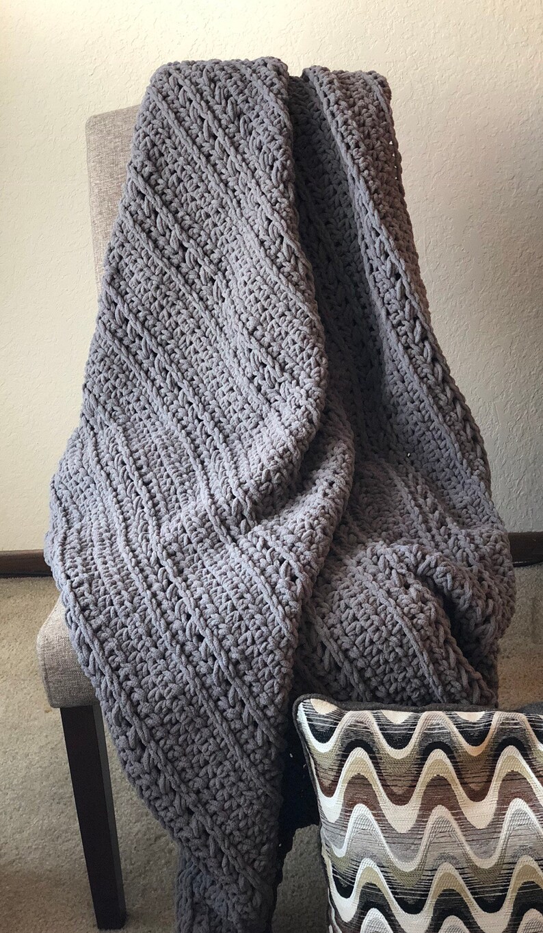 May include: A large, gray, hand-crocheted blanket draped over a gray upholstered chair. The blanket has a thick, textured weave. A decorative pillow with a wavy pattern in shades of brown, gray, and white is visible.